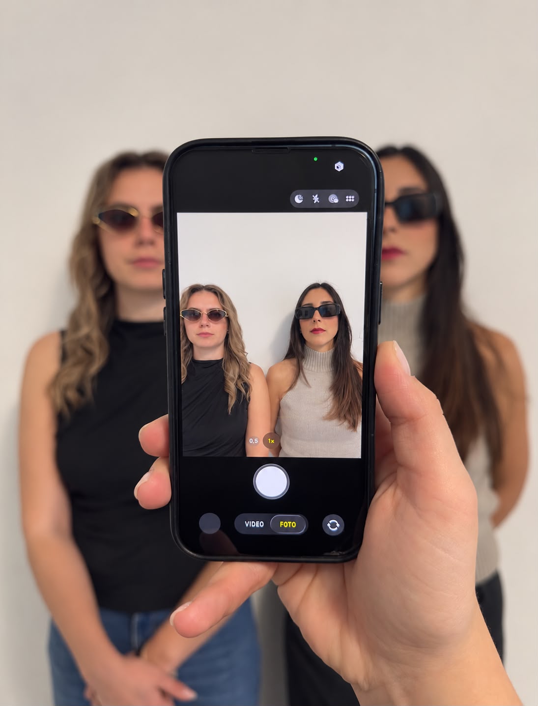 Una mano inquadra con lo smartphone Alessandra Ajdini e Martina Bianchini, in piedi contro una parete chiara con gli occhiali da sole